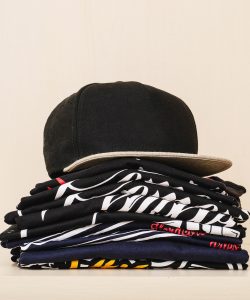 Pile-of-brand-new-t-shirts,-dark-tones,-with-print-and-a-black-straight-flap-cap-over-the-pile