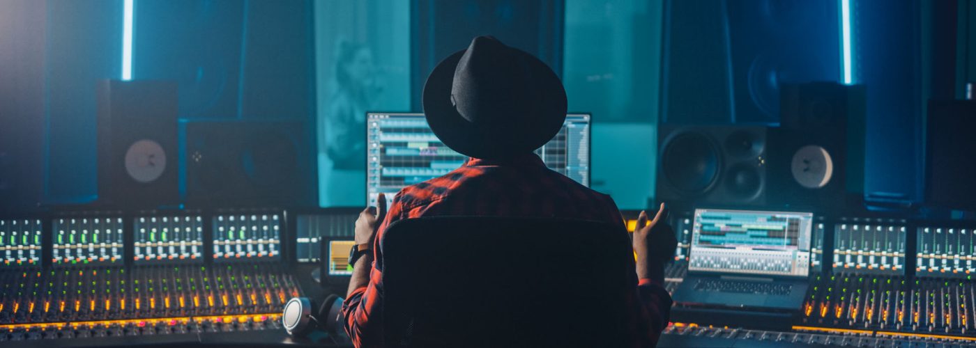 Producer, Audio Engineer Uses Control Desk for Recording New Album Track in Music Record Studio, in the Soundproof Room Musician, Artist, Performer Sings a Song from New Album. Back View